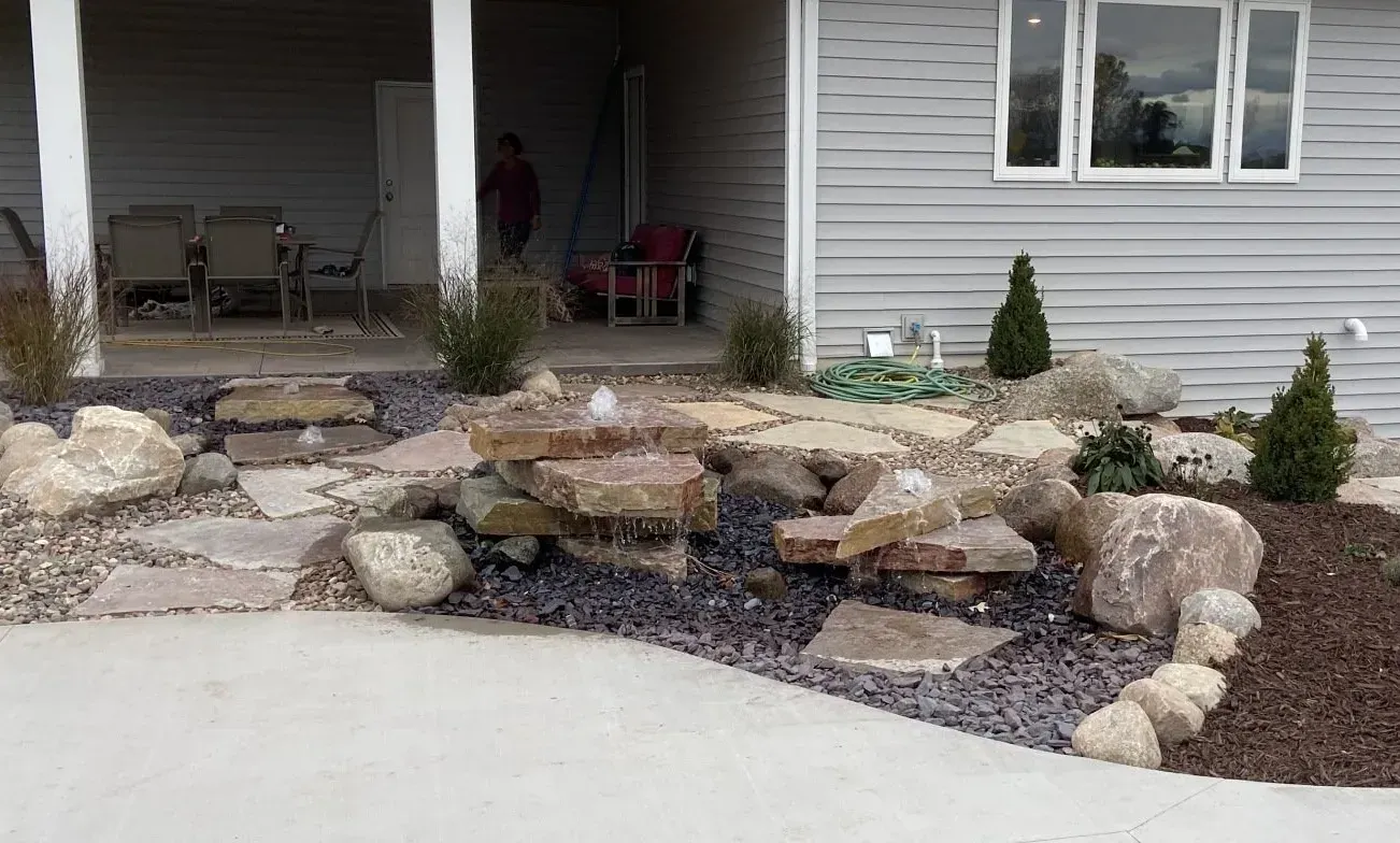 Boulder water feature and stone bed beside a concrete patio and home