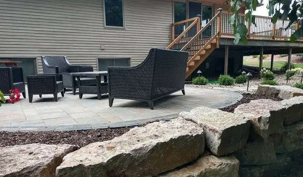 Wicker patio chair on a stone patio edged with boulders below a deck stairway